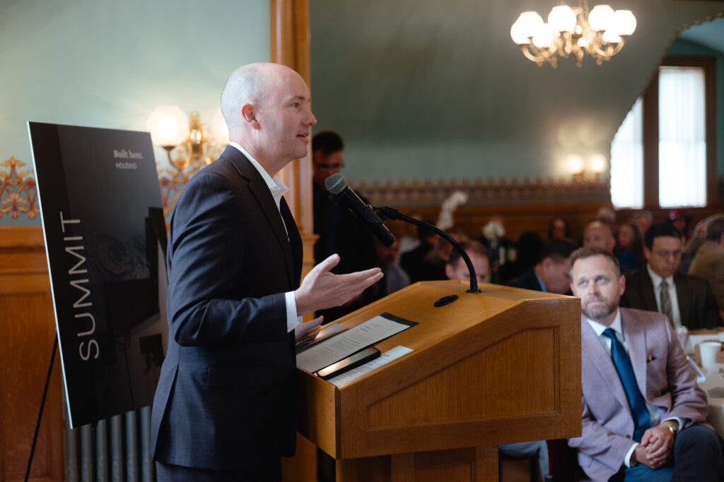Utah Gov. Spencer Cox stands behind a wood podium addressing a medium-sized crowd.